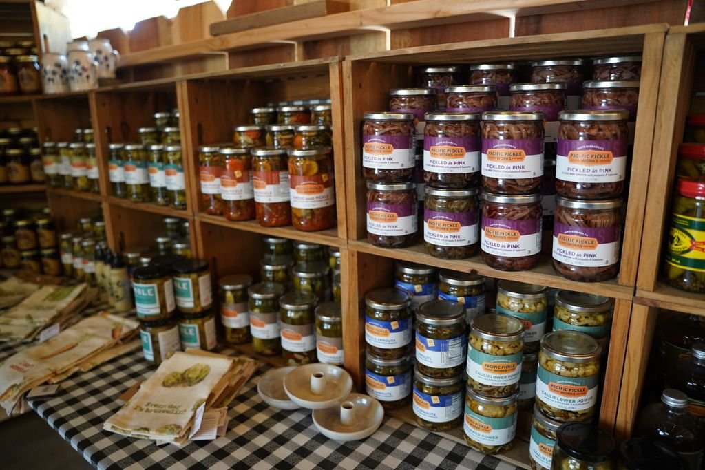 An assortment of jars filled with pickled vegetables displayed on wooden shelves in a rustic shop setting.